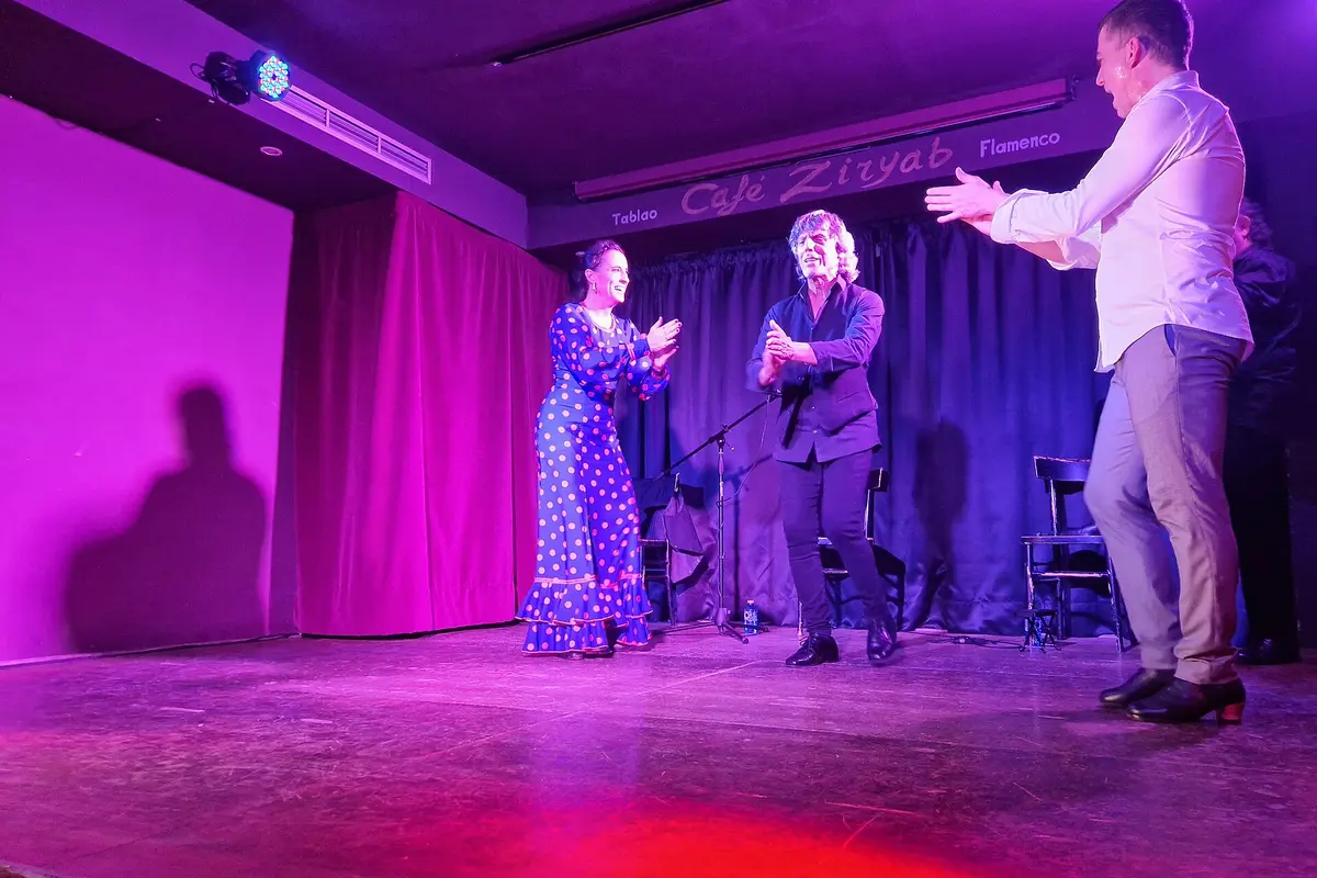Uma bailaora num vestido azul de bolinhas, um guitarrista e um segundo homem a bater palmas num pequeno palco sob luzes cor-de-rosa e azuis